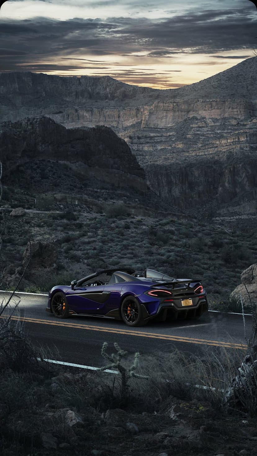 Purple McLaren convertible racing down a winding desert road at sunset between rugged canyon rock walls.