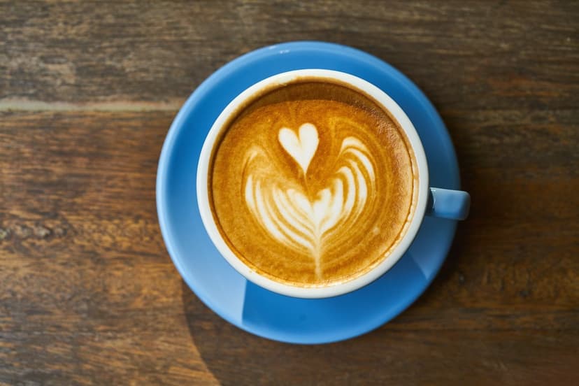 Heart-shaped foam latte art in a white cup on a blue saucer atop a dark wood grain table from an overhead view.