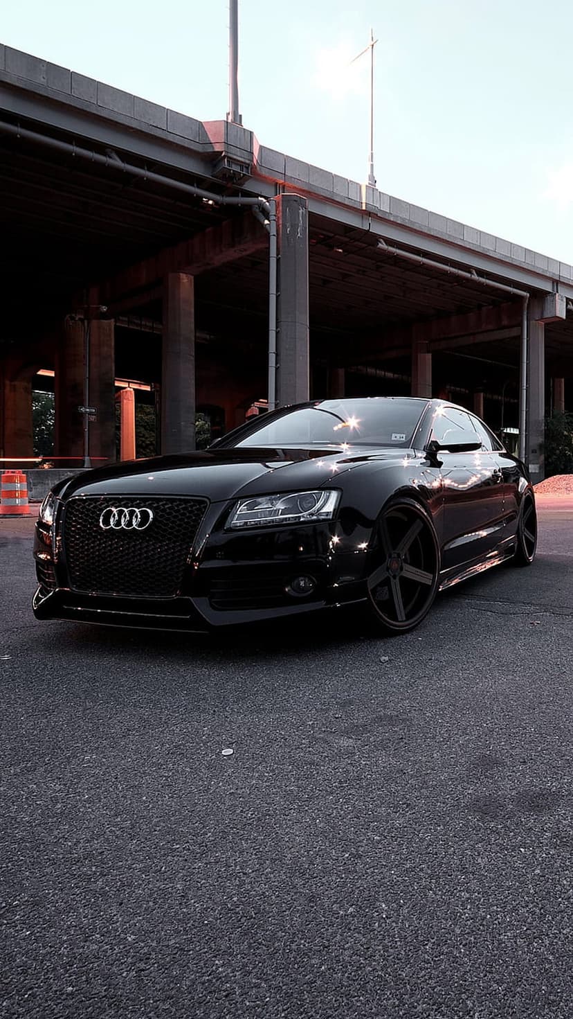 Black Audi coupe parked under a concrete overpass during dusk, featuring aggressive styling and custom wheels.