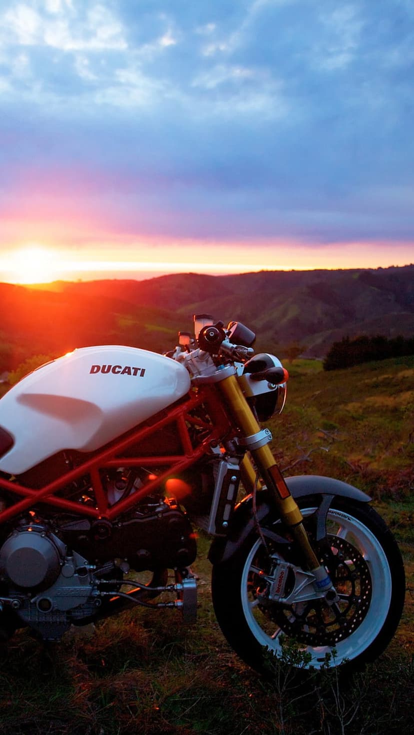 White Ducati motorcycle parked on a grassy ridge against a pink and orange sunset sky with distant mountains.