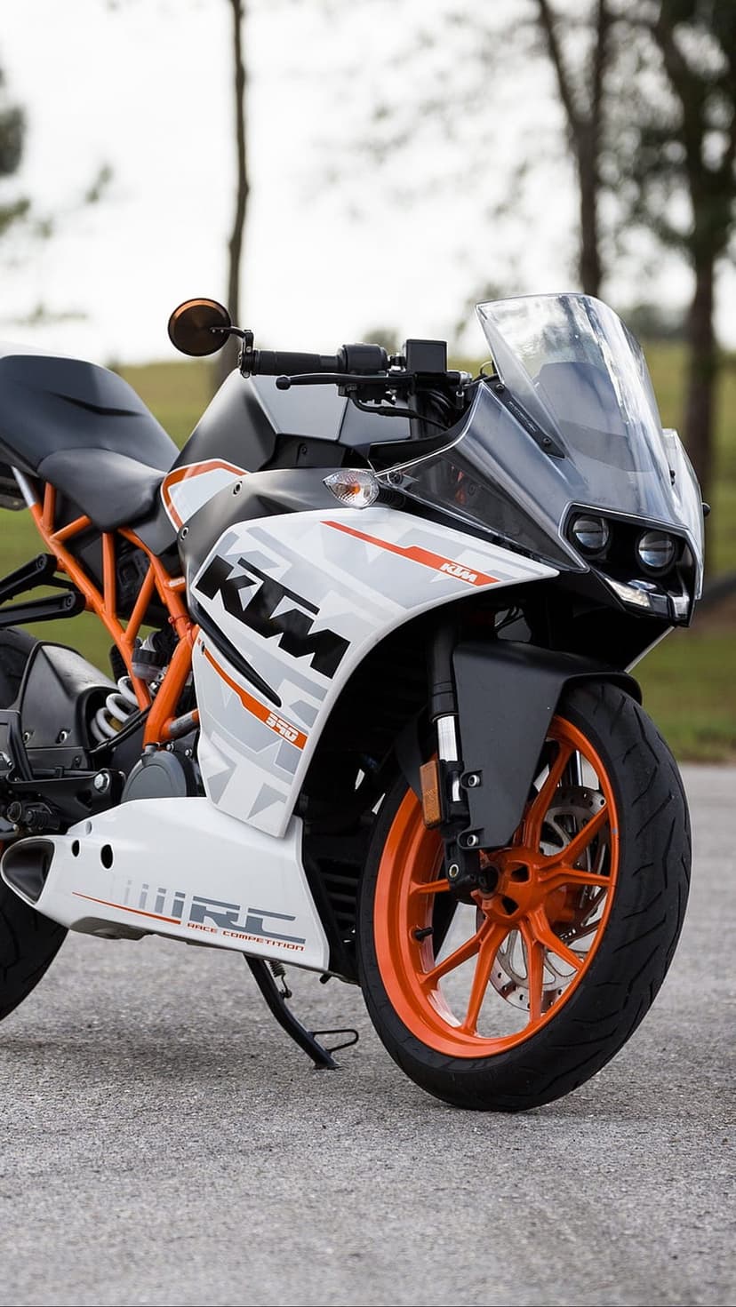 White KTM RC 390 motorcycle with bright orange wheels parked on a paved surface with a blurred forest background.