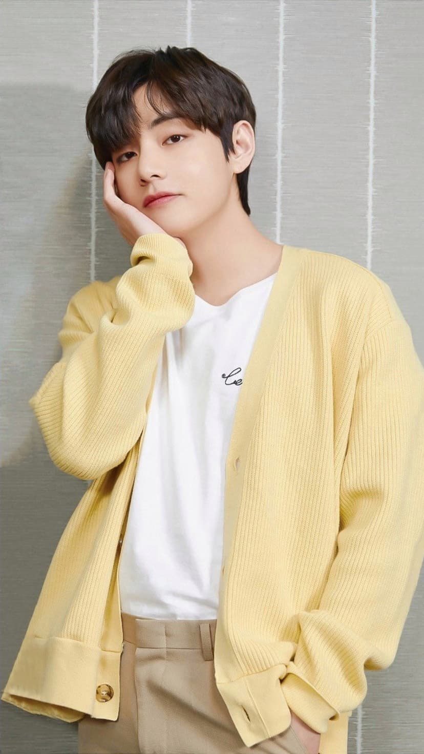 Kim Taehyung of BTS wearing a yellow cardigan with his hand on his cheek against a textured gray wall background.