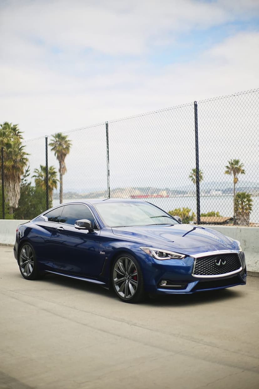 Blue Infiniti Q60 coupe driving on a coastal road past palm trees and a sparkling ocean under bright sunlight.