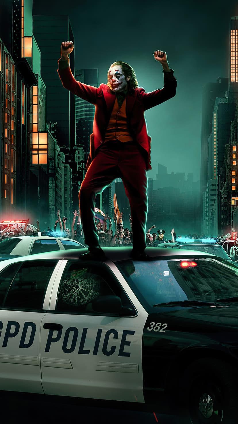 Joker in red suit stands on a police car with raised arms amidst a cheering crowd in a dark city night.