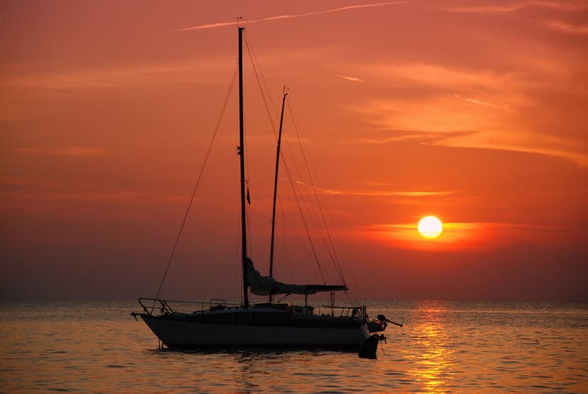 Sailboat silhouette rests on calm ocean waters under a vibrant orange sunset with glowing sun on the horizon.