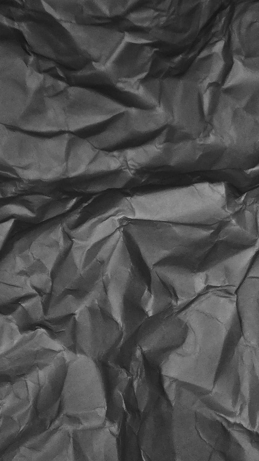 Charcoal gray crumpled paper with sharp creases, deep shadows, and subtle highlights in a vertical close-up.