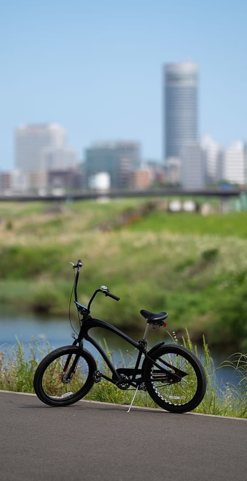 Black cruiser bicycle stands on a paved trail beside a river with a blurred city skyline and bridge behind it.