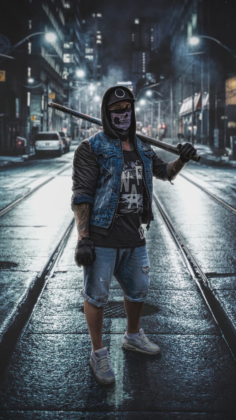 A person in a skull mask and denim jacket holds a long staff while standing on dark city train tracks at night.