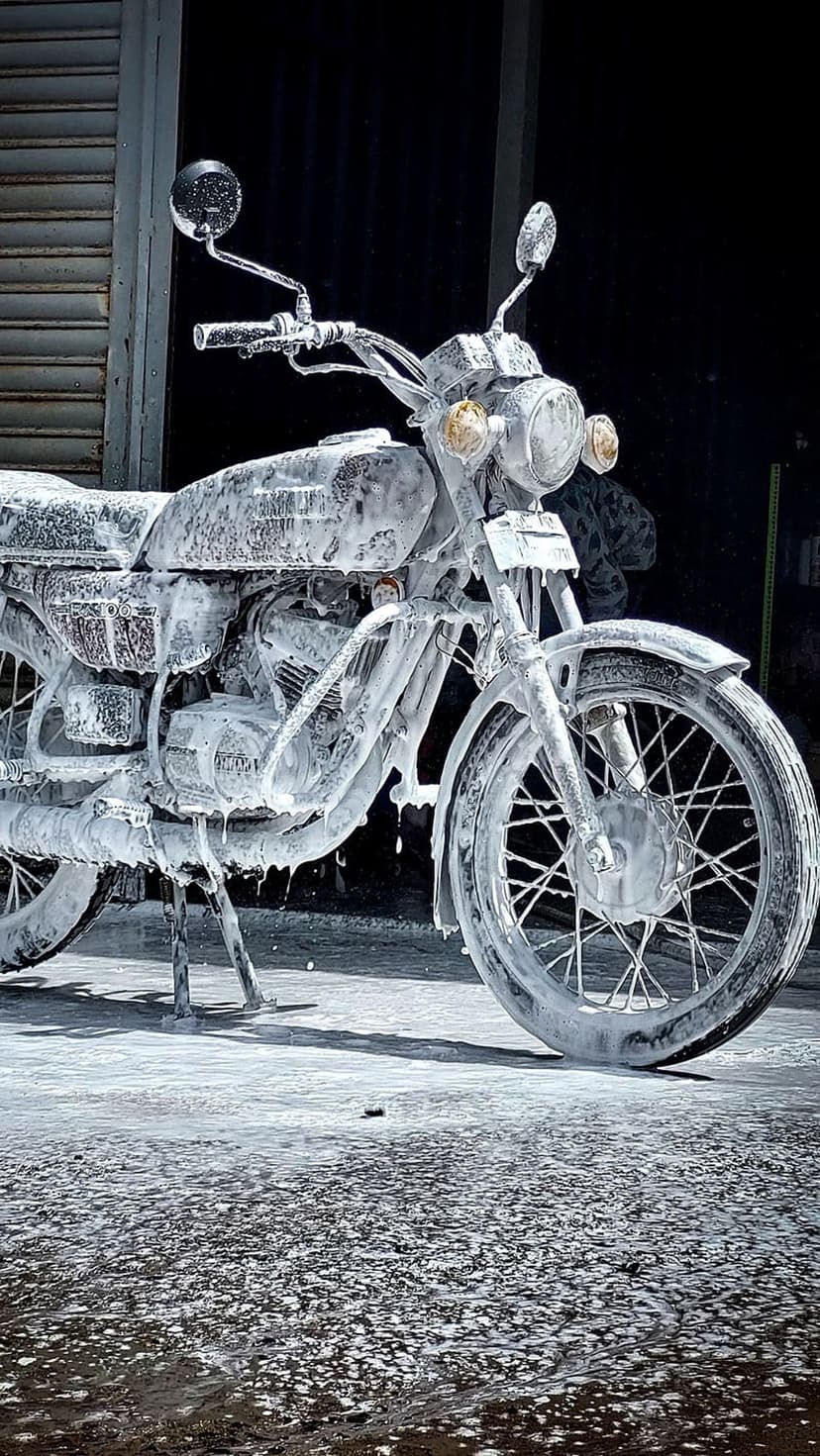 Vintage motorcycle covered in thick white foam suds with visible handlebars and spokes against a dark background.