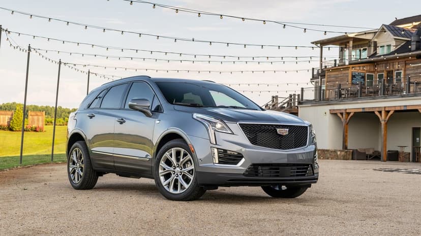 Gray Cadillac SUV on gravel with a rustic wooden building and string lights under a cloudy evening sky.