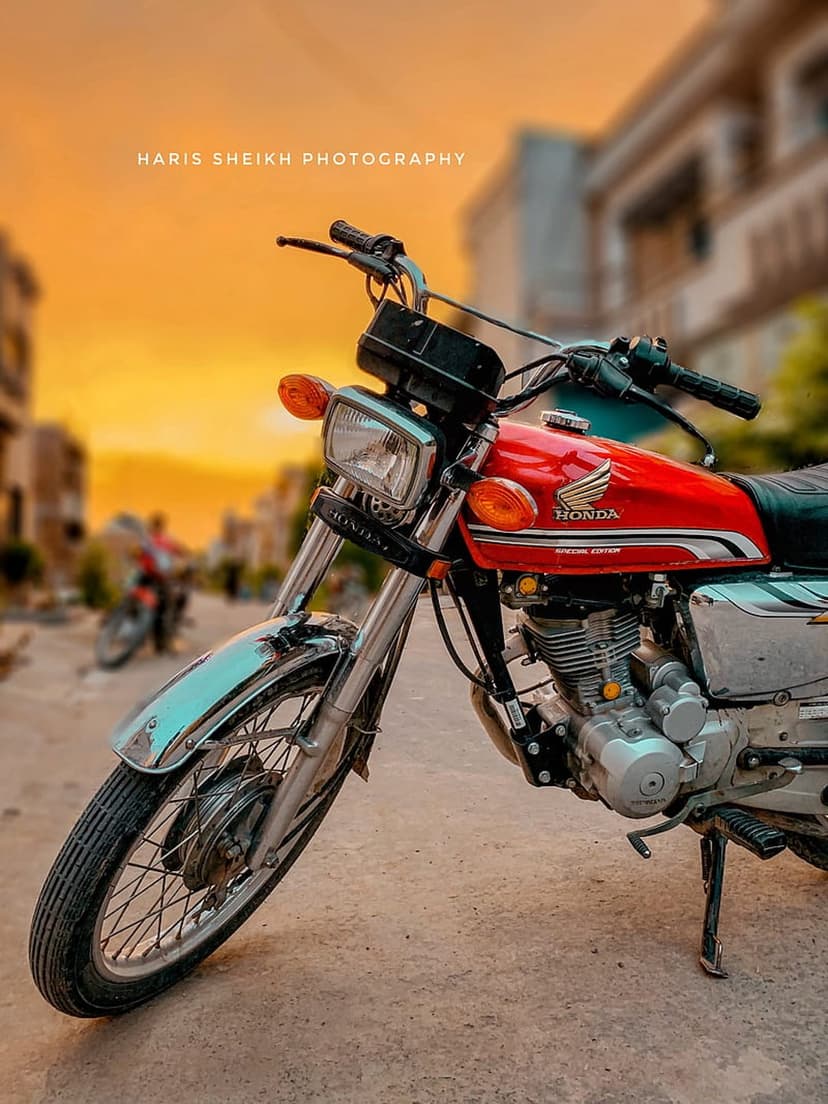 Red Honda motorcycle with chrome engine parts parked before a blurred city skyline under an orange sunset sky.