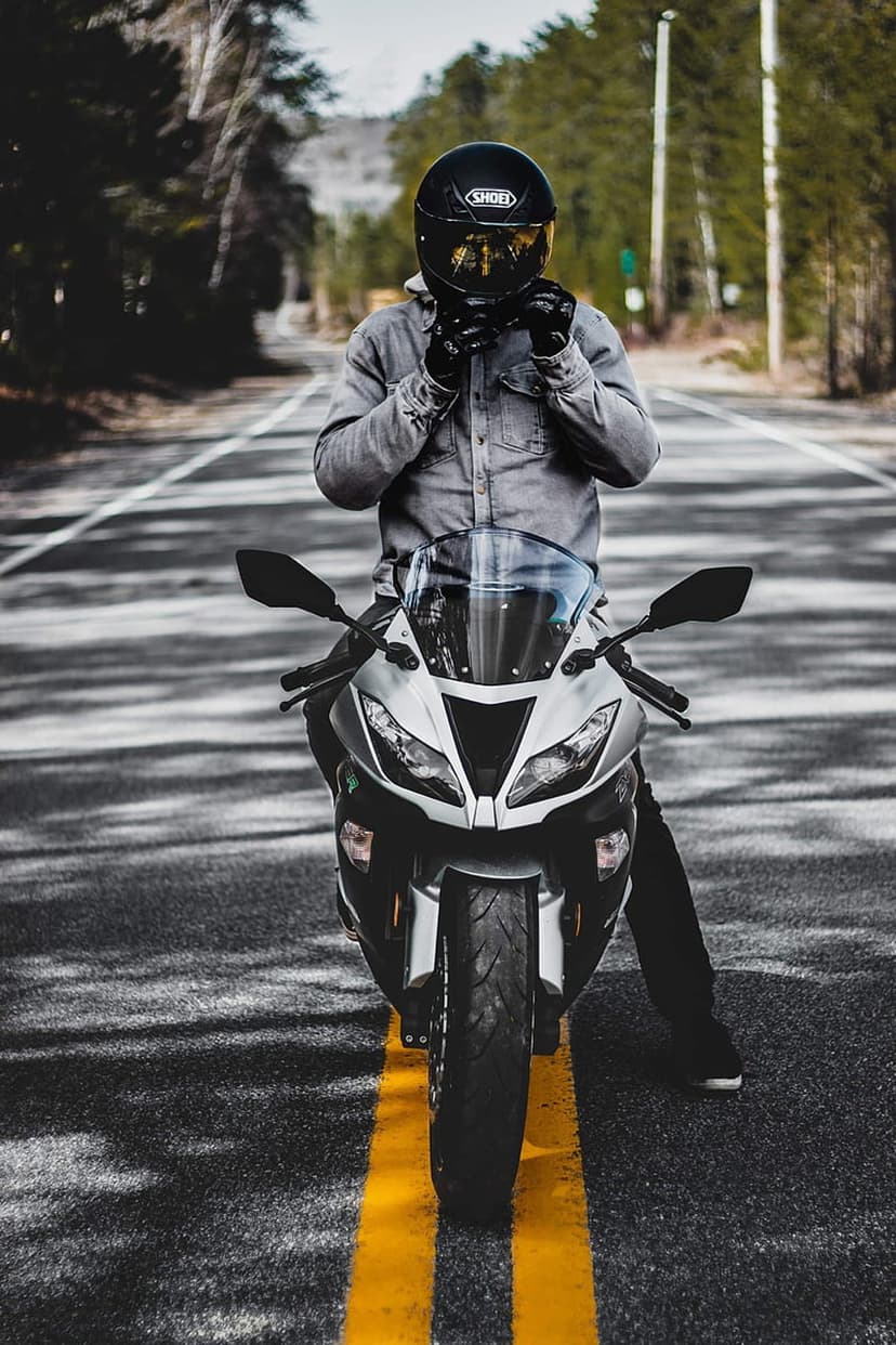 Motorcyclist on a silver sportbike adjusts a black helmet with a gold visor on a forest road with yellow lines.