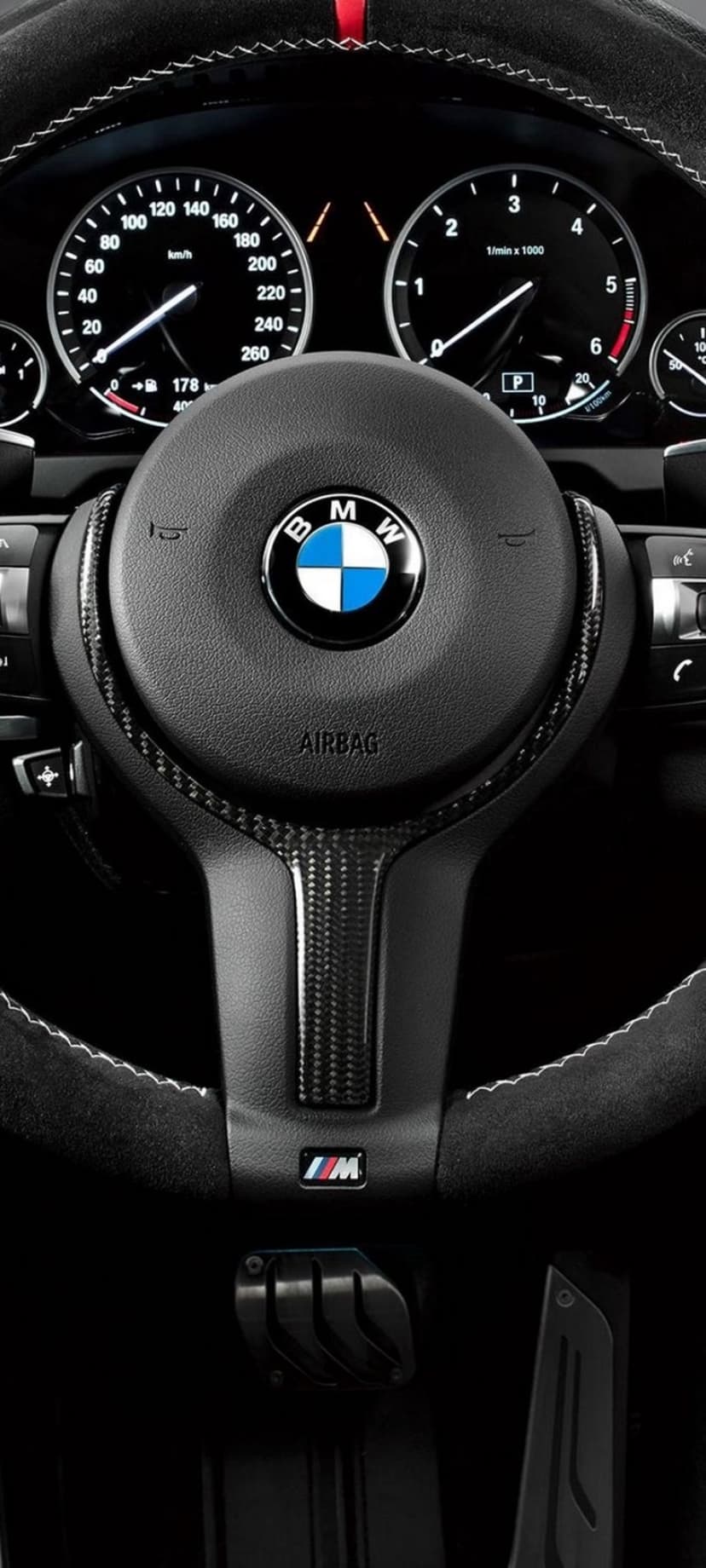 BMW M steering wheel with carbon fiber trim and a tricolor logo positioned in front of a lit vehicle dashboard.