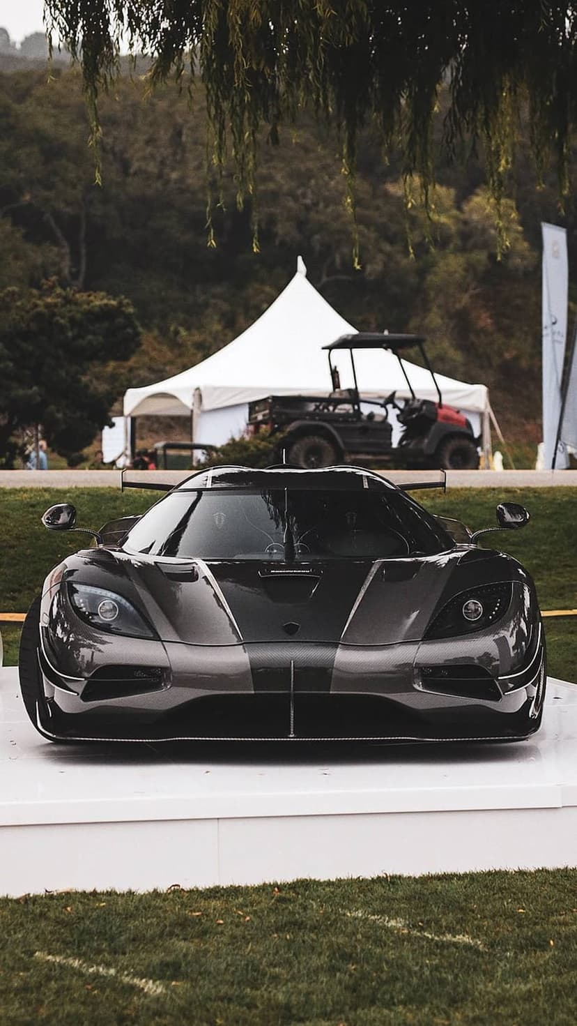 Dark grey Koenigsegg supercar with racing stripes sits on a white display platform amidst green trees and grass.