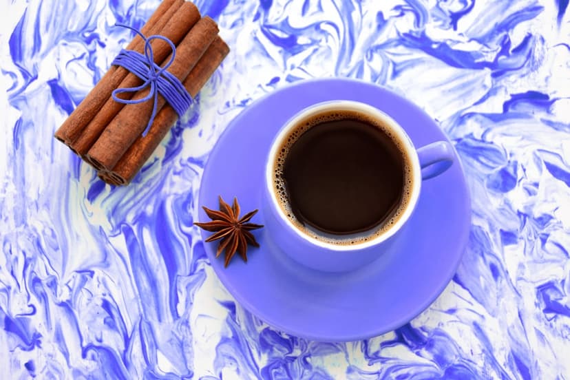 Cup of black coffee on a purple saucer next to cinnamon sticks and star anise over blue marble stone.