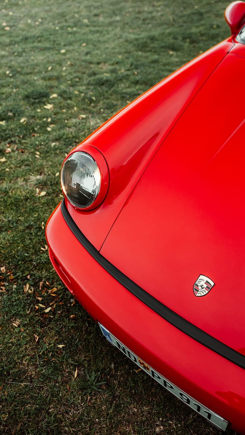Red Porsche 911 front hood and round headlight sitting on green grass with scattered brown fallen leaves.