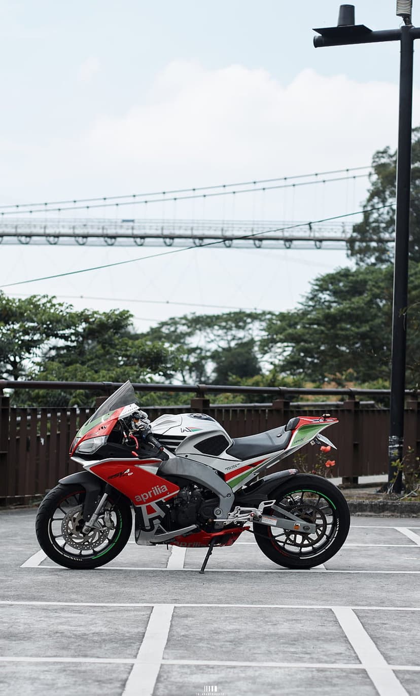 Red and white Aprilia sportbike parked on concrete with a large suspension bridge and green trees in the background.