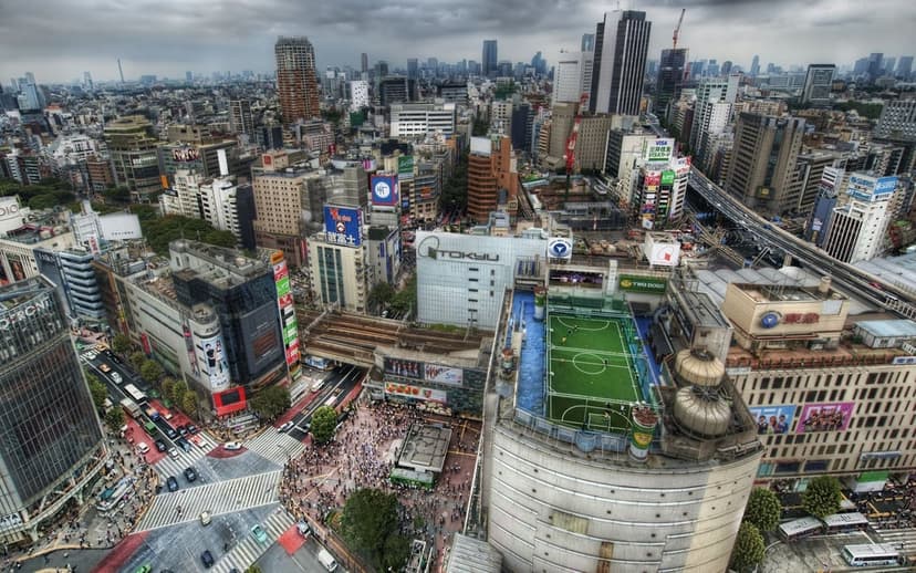Rooftop soccer field surrounded by tall gray buildings and busy city streets under an overcast sky in Tokyo.
