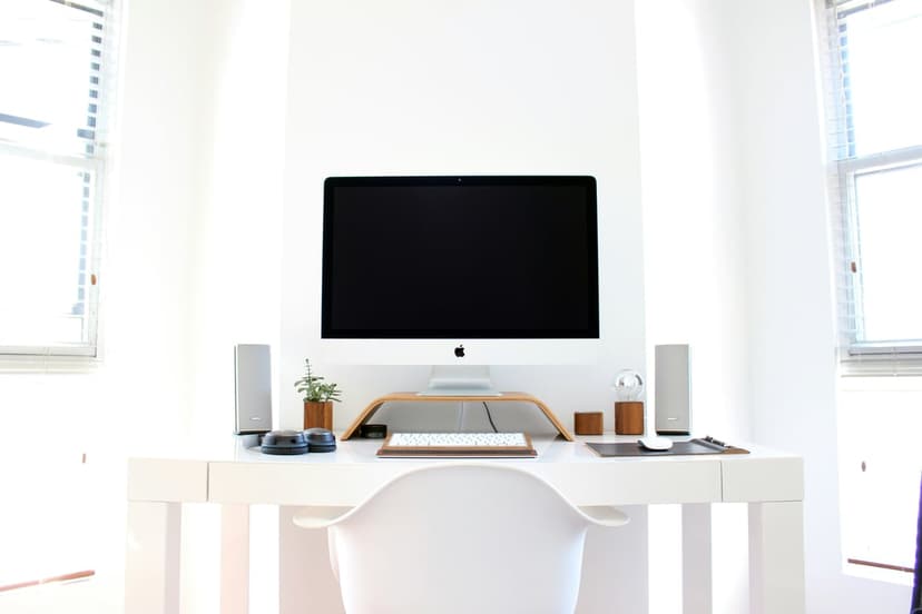 White desk setup with an Apple iMac, silver speakers, and a small plant illuminated by natural sunlight.