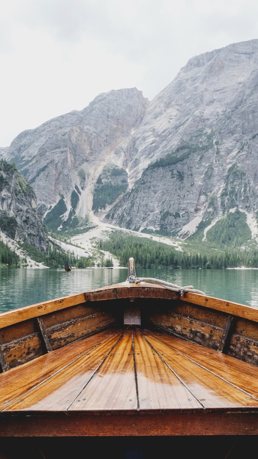 Wooden rowboat on a still mountain lake reflecting rocky peaks and green pine forests under an overcast sky.
