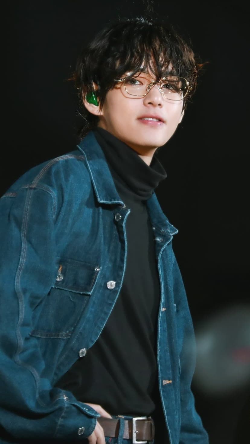 Kim Taehyung of BTS smiles wearing a dark denim jacket, black turtleneck, gold glasses, and green earbuds.