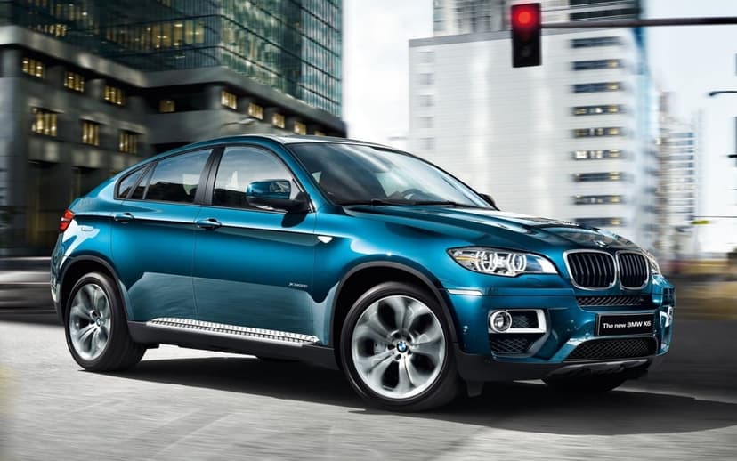 Blue BMW X6 SUV driving past skyscrapers with motion blur on wheels and a red traffic light in the city.