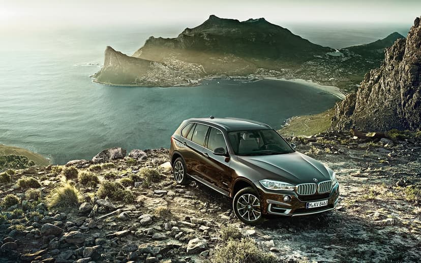 Dark brown BMW SUV on a rocky cliff overlooking a turquoise bay and misty mountains at sunset.