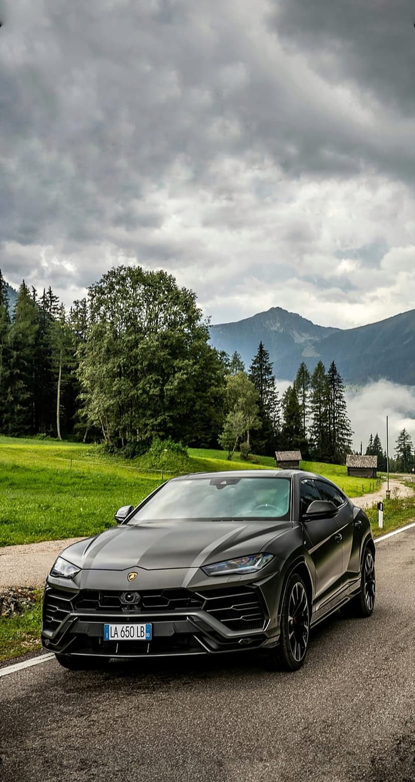 Dark grey Lamborghini Urus SUV on a winding mountain road with green forests and cloudy peaks in the background.