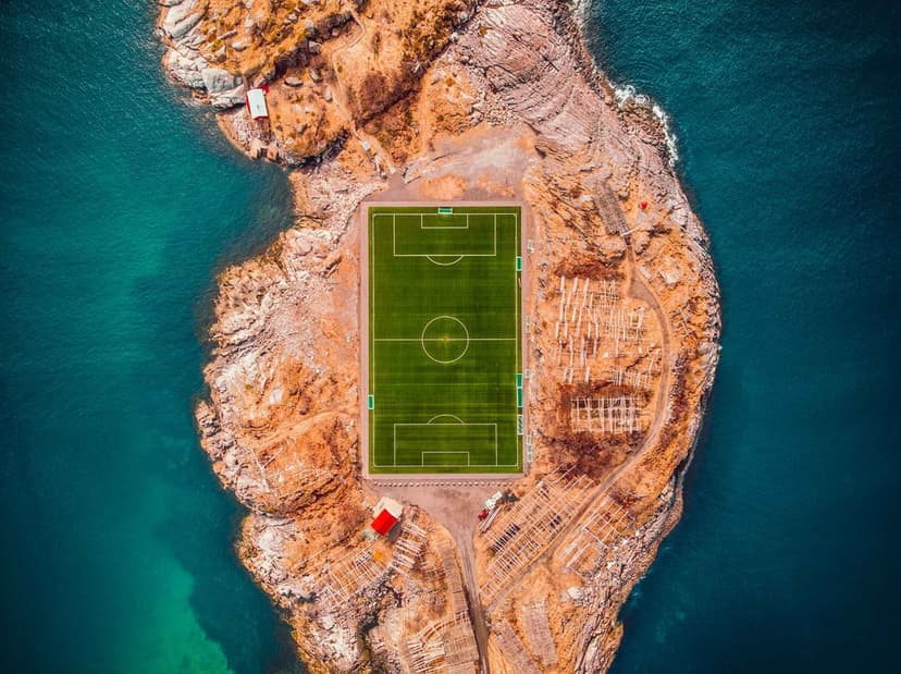 Vibrant green football pitch centered on a jagged rocky island surrounded by swirling turquoise ocean water.
