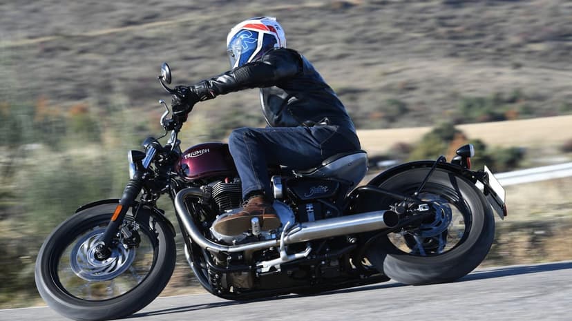 Triumph Bonneville Bobber leaning into a curve on a road with a rider in leather and a colorful helmet.