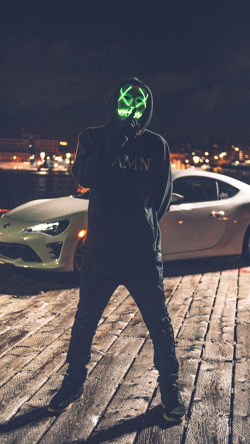 Masked person with glowing green X eyes gestures for silence on a wooden pier near a white sports car at night.