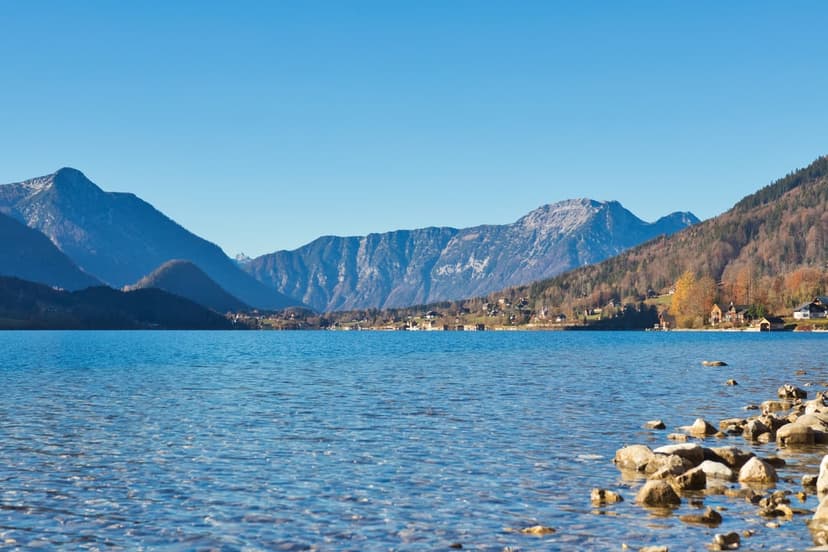 Snow-dusted mountain peaks reflect in a calm blue lake beside a small village surrounded by autumn trees.