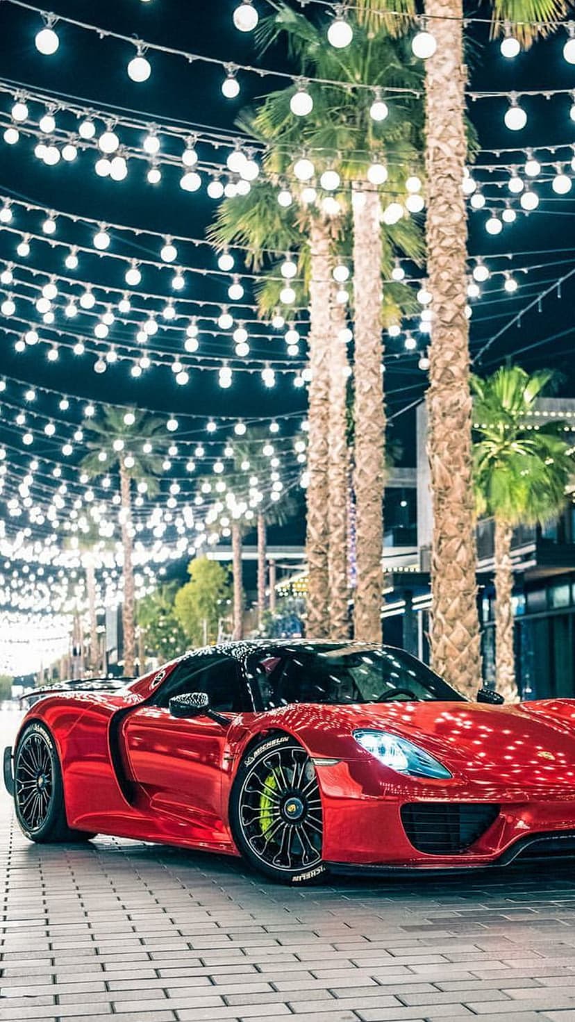 Red sports car parked on a brick path under glowing string lights and tropical palm trees during a summer night.