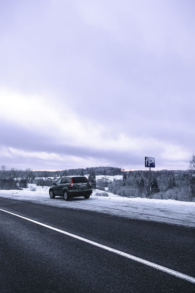 Dark SUV parked on a snowy road shoulder next to a blue parking sign with hills and a cloudy winter sky.