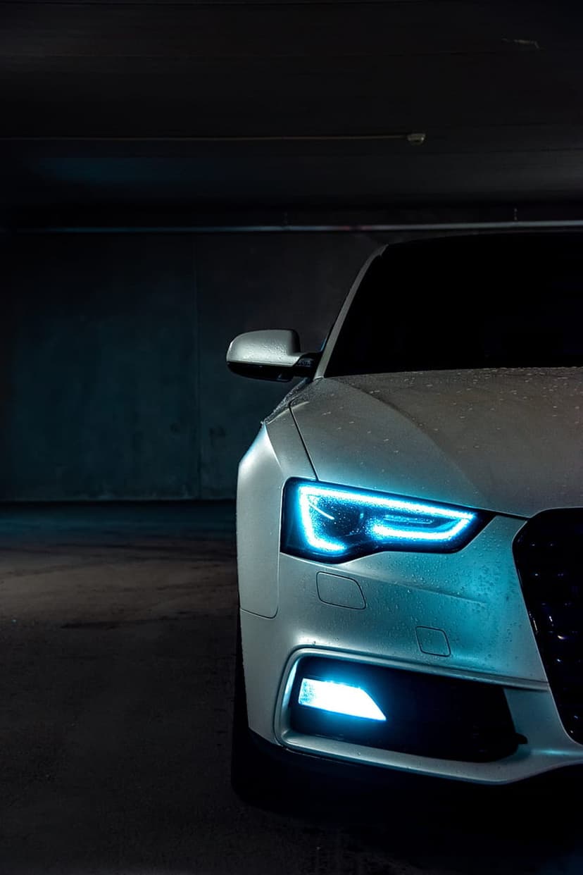 White Audi car front with glowing blue LED headlights and water droplets in a dark concrete parking garage.