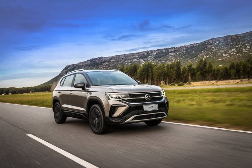 Volkswagen Taos SUV driving on a highway with a rocky mountain and green forest under a blue sky background.