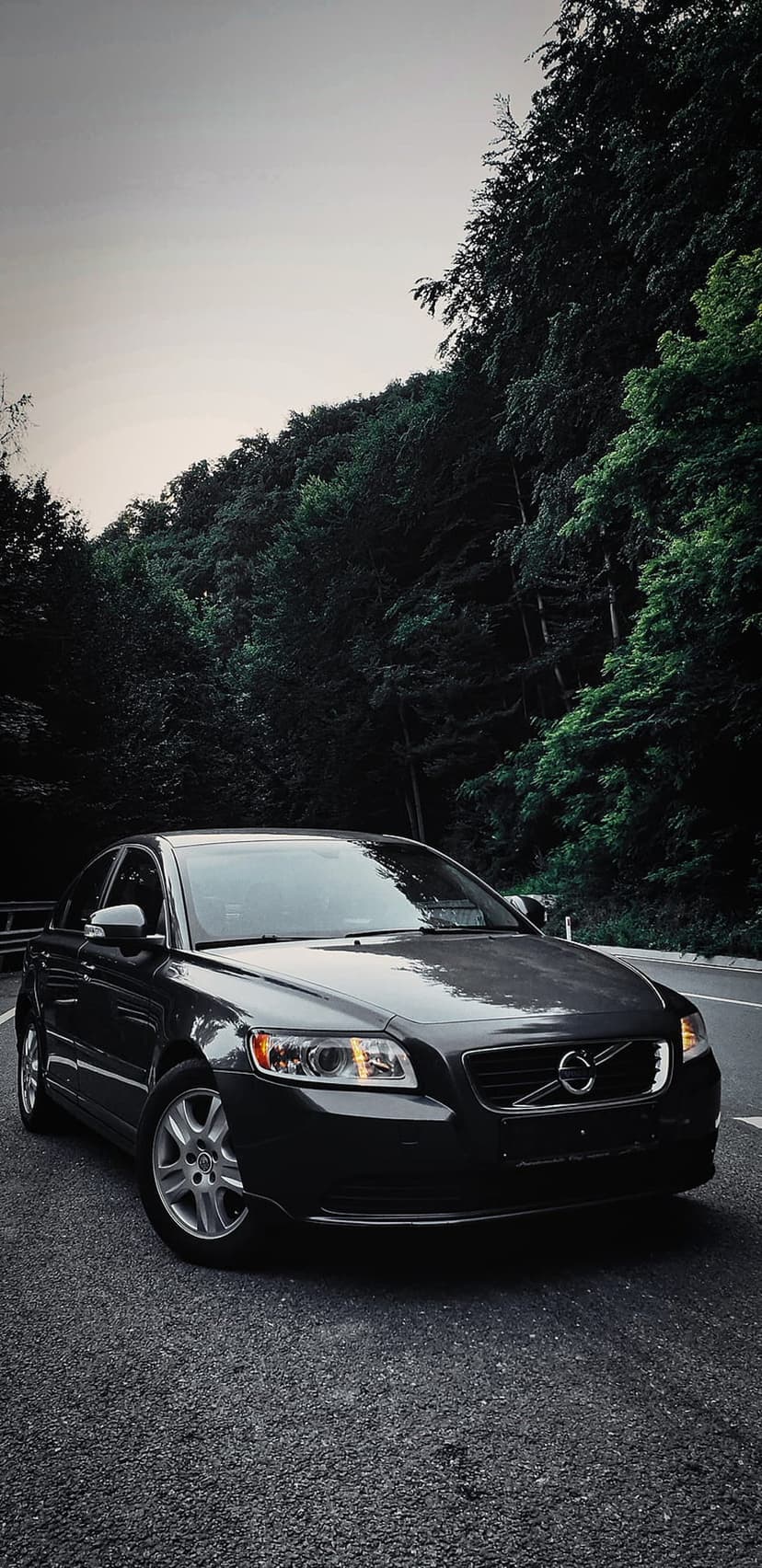 Volvo S40 sedan with glowing headlights parked on a textured asphalt road surrounded by dense green forest.