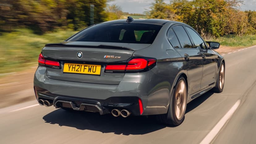 Grey BMW M5 CS sports sedan driving fast on a sunlit road with gold wheels and carbon fiber details.