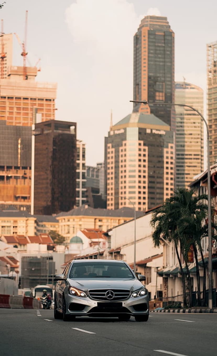 Silver Mercedes E-Class sedan driving on a city street between skyscrapers under warm evening sunlight.