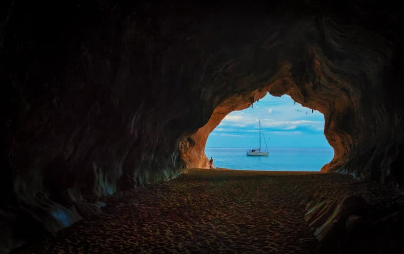 Sailboat anchored on a bright blue sea as seen from the dark, craggy interior of a sandy coastal limestone cave.