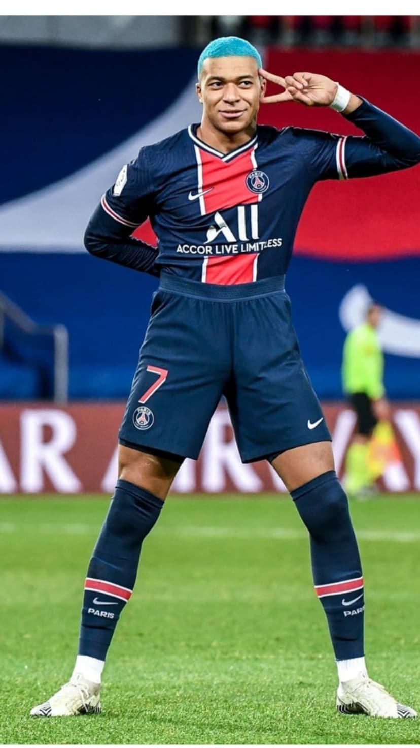 Kylian Mbappé with blue hair celebrates a goal in a navy PSG kit during a match on a lush green grass field.