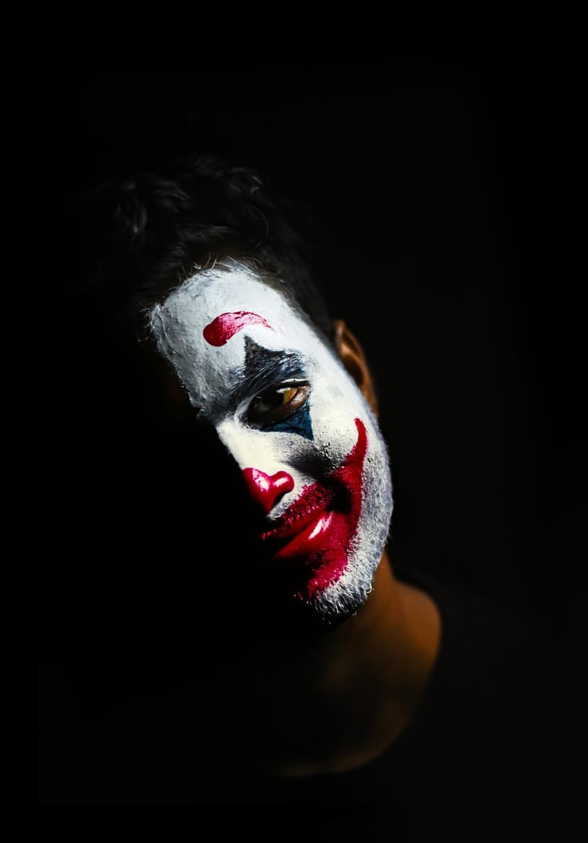Man with white and red clown makeup stares intensely through dramatic shadows in a close-up vertical portrait.
