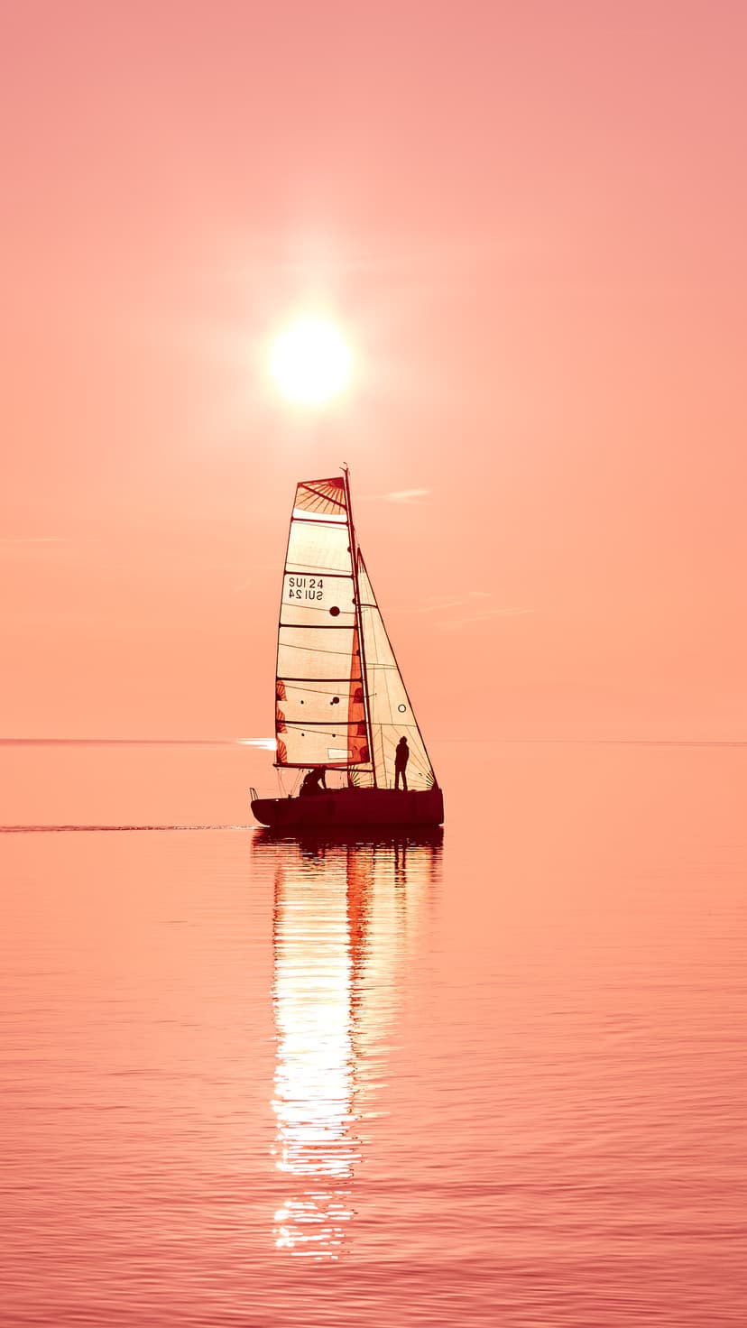 Sailboat silhouette rests on a calm pink lake during sunrise with soft peach clouds reflecting in the still water.