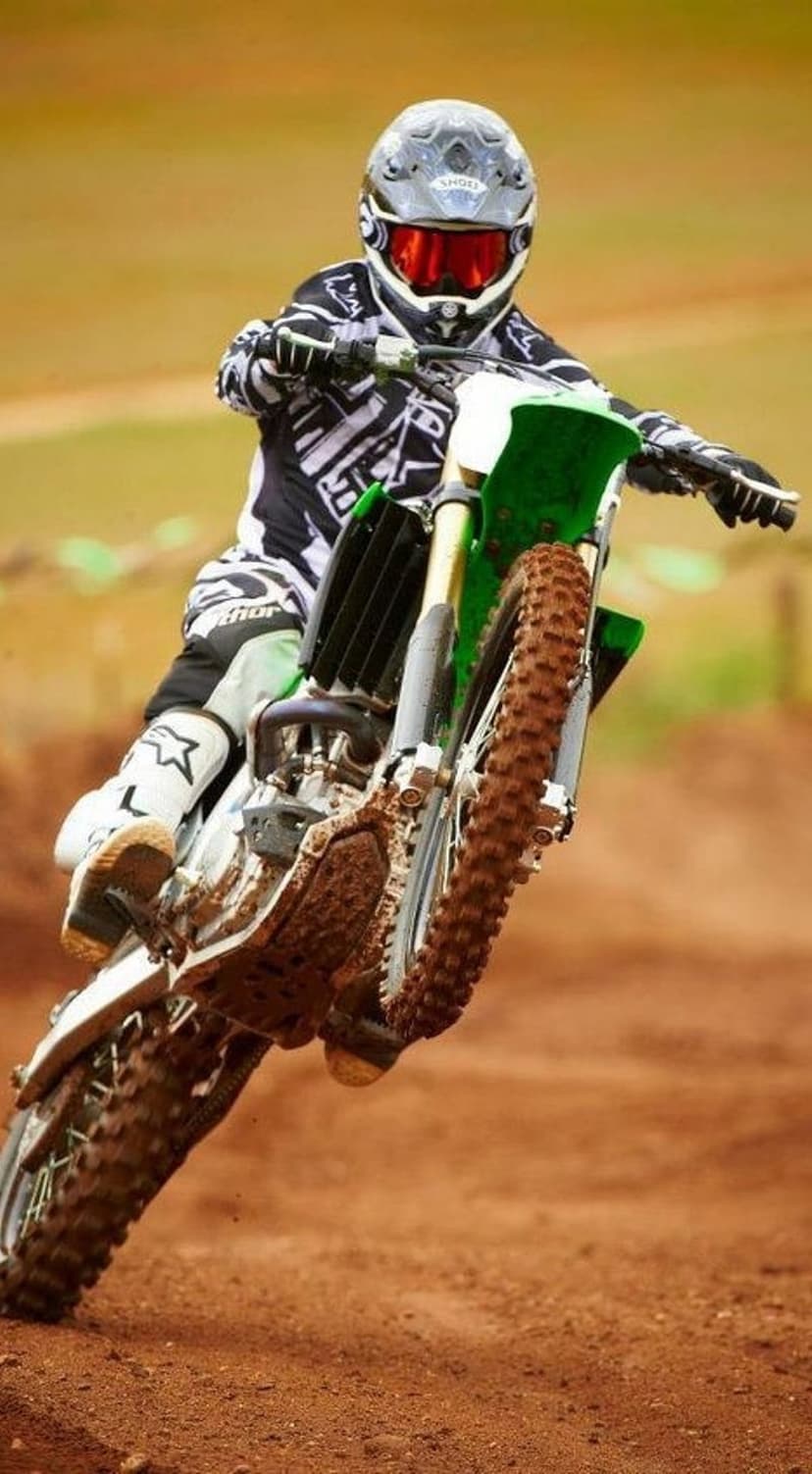 Motocross rider wearing red goggles performs a high jump on a motorcycle with mud spraying from the back tire.