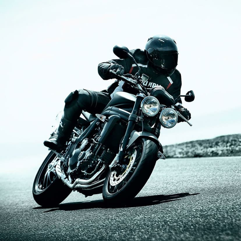 Black Triumph motorcycle leaning into a sharp turn on a paved road with a rider wearing full black leather gear.