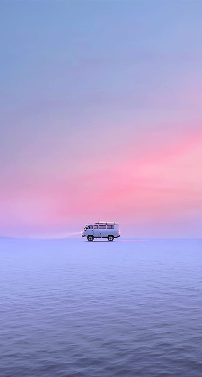 White van with glowing headlights driving across a reflective water surface under a pink and purple sunset sky.