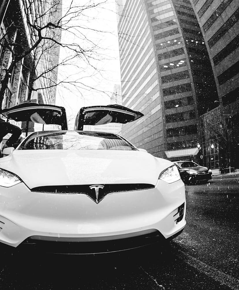 White Tesla Model X with open Falcon Wing doors on a snowy city street framed by tall glass skyscrapers.