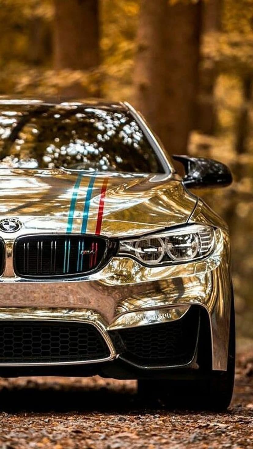 Gold chrome BMW M sports car parked in a sunlit autumn forest with blurred orange leaves in the background.