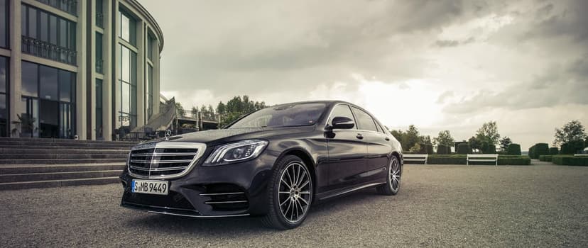Black Mercedes-Benz S-Class sedan with raindrops on a gravel path near a majestic curved building.