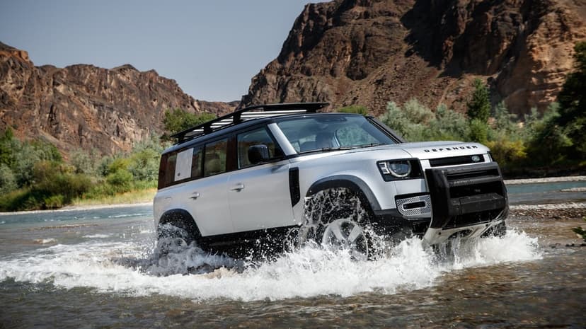 Silver Land Rover Defender SUV splashing through a river in a mountainous desert canyon landscape.